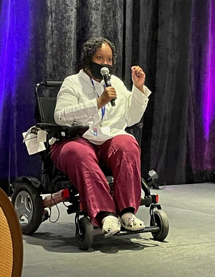A photo of a young black woman in a wheelchair, speaking on a microphone to a group, with a mask on her face.