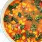 A vibrant vegetable soup is pictured, featuring chunks of yellow squash, diced tomatoes, leafy greens, and white beans in a broth. The ingredients are dispersed evenly, creating a colorful and hearty appearance.
