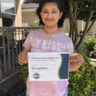 Permalink to How Toastmasters Has Helped Me A person stands outdoors, smiling and holding a Toastmasters certificate for completing Level 2 Pathways. They wear a pink tie-dye shirt and blue jeans, with a building and greenery in the background.
