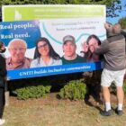 Permalink to Surrey council moves Harmony project forward Three people are adjusting a large sign in front of a development proposal notice. The sign features diverse individuals and reads Real people. Real need. and UNITI builds inclusive communities. Bushes are in the background.