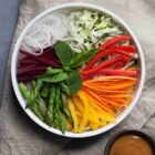 Permalink to Noodle Bowl with Rainbow Veggies & Peanut Sauce A colorful salad in a white bowl with sliced beets, asparagus, onions, cabbage, red and yellow bell peppers, and carrots, topped with mint leaves. A small bowl of dressing is beside the salad on a textured cloth.