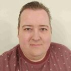 Permalink to Full-Time Freedom A person with short hair is wearing a maroon shirt with small white geometric patterns. They are against a plain light-colored background, looking directly at the camera with a neutral expression.