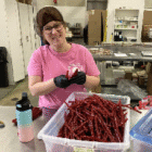 A person wearing a pink shirt, glasses, and a hairnet is smiling while packaging red licorice in a factory setting. They are holding a small package of licorice, and there are large containers filled with the candy on the table.