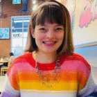 Permalink to My Happiness Project A person smiling, wearing a colorful striped sweater and a beaded necklace, is seated indoors at a café. The background features brick walls, art on the walls, and another person sitting at a table.