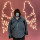 Permalink to My Story About Canva A person wearing a mask and a blue jacket stands in front of a pink wall with illuminated neon angel wings.