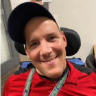 Permalink to Being Valued A man wearing a blue baseball cap and a red shirt smiles at the camera. He is seated in a wheelchair and has a lanyard around his neck with Seattle Seahawks written on it. The background shows an office environment.