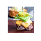 Permalink to Keto Big Mac Bites A juicy cheeseburger slider topped with melted cheese, lettuce, and two pickle slices. It is secured with a skewer and displayed on a wooden surface. A blurred dish is in the background.