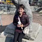 Permalink to My Five Things A person in a dark jacket sits on a stone next to a bear sculpture, holding a coffee cup. They are outside, with buildings and a fence in the background.